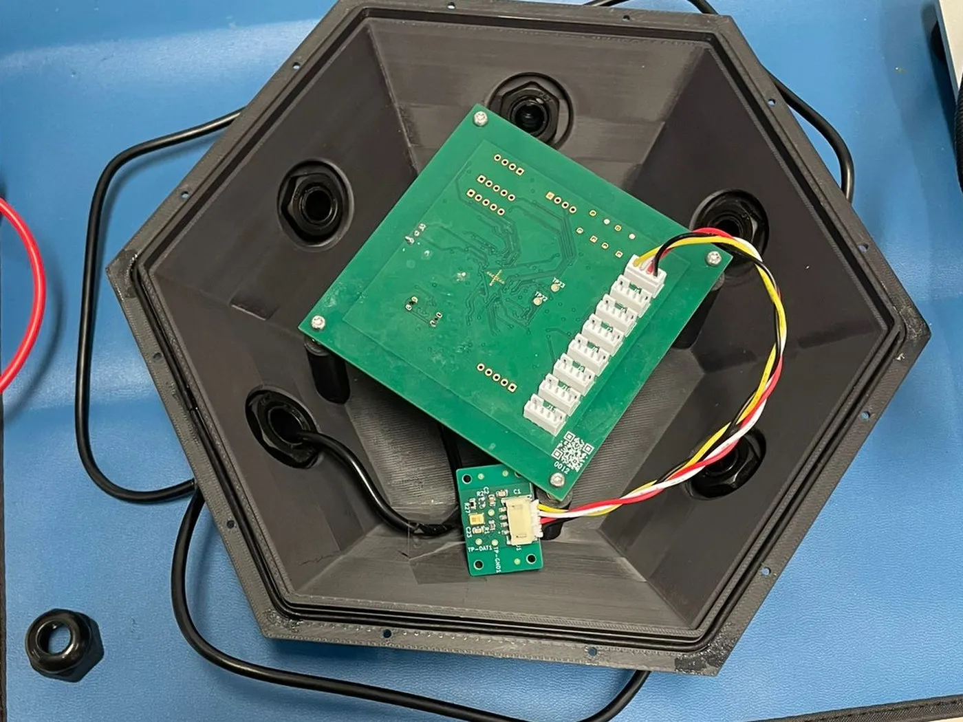 Ein geöffnetes Knoten-Basisgehäuse auf einer ESD-Matte mit der Hauptplatine und einer kleinen Trägerplatine darin, daneben ein Multimeter