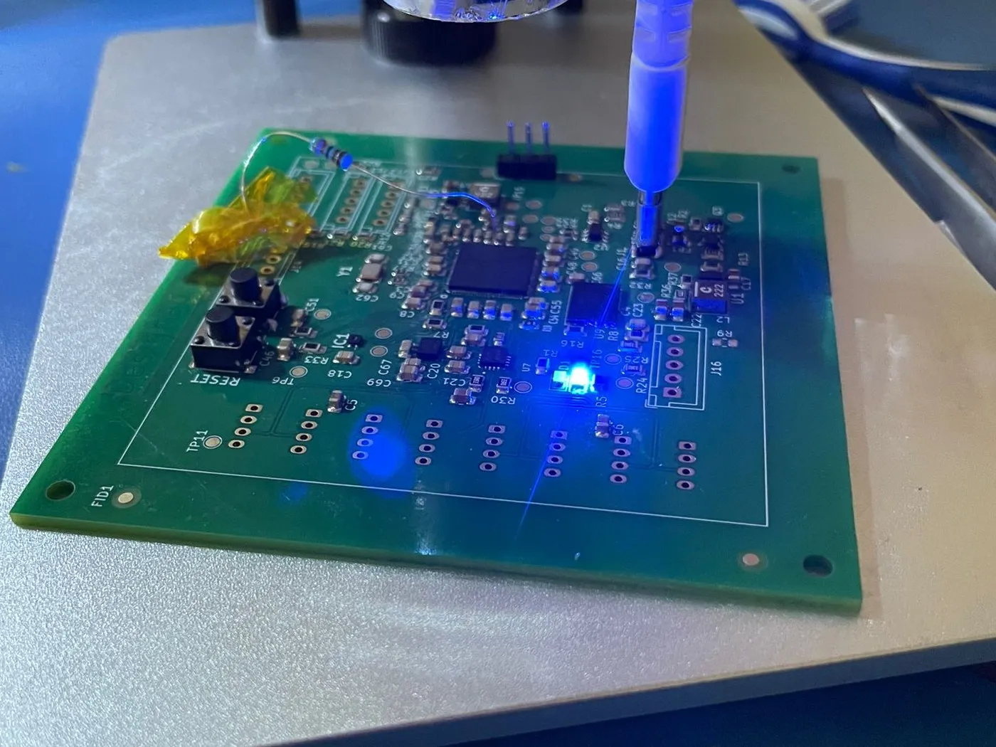 Dieselbe Platine auf dem Mikroskoptisch während der Inbetriebnahme, eine Prüfspitze auf einem Pad, eine Status-LED leuchtet, dazu ein Fliegdraht und Kaptonband