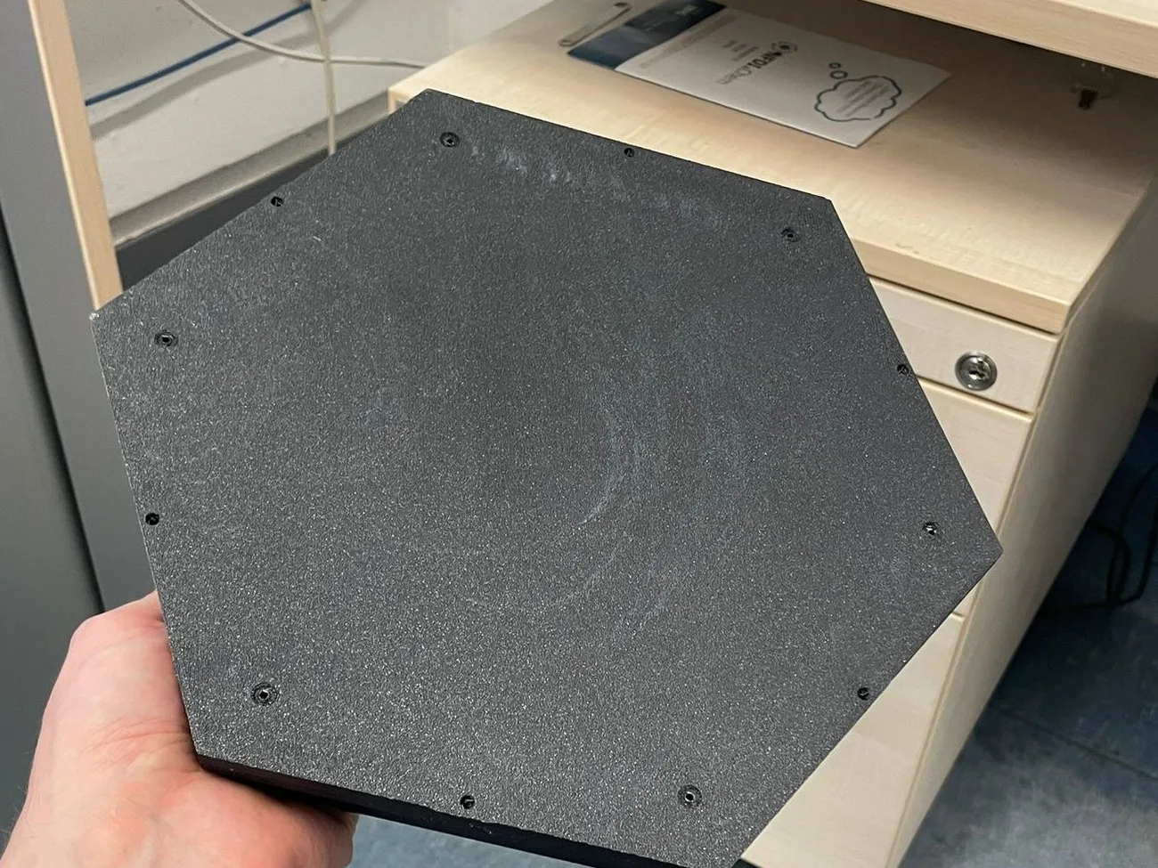 Eine matt schwarze, 3D-gedruckte sechseckige Deckplatte, in einer Hand in der Werkstatt gehalten